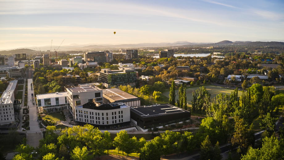 An aerial view of the Australian National University campus, with a hot air balloon the Canberra Civic centre and Lake Burley Griffin in the background.