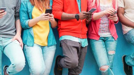 A group of young people lean against a wall. They are wearing mostly bright outfits and scrolling on their phones.