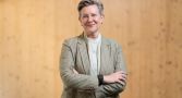 Professor Joan Leach stands with her arms crossed, smiling. She is wearing a gray blazer.