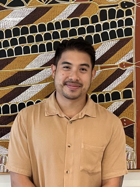 Man with short black hair standing in front of painting wearing light brown shirt and smiling