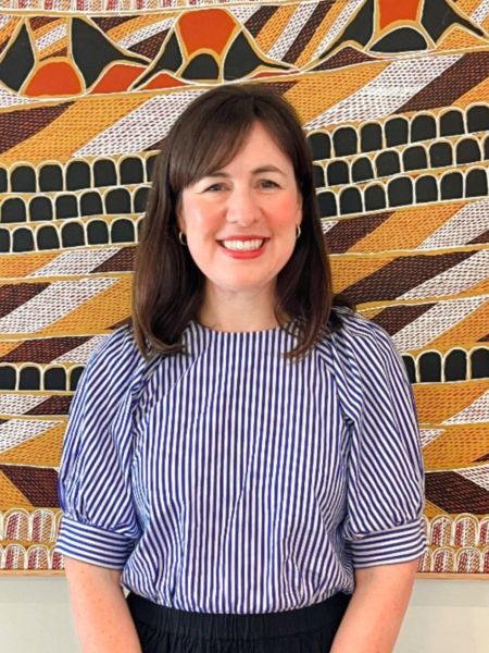 A person standing in front of a painting smiling wearing a stripy blue shirt